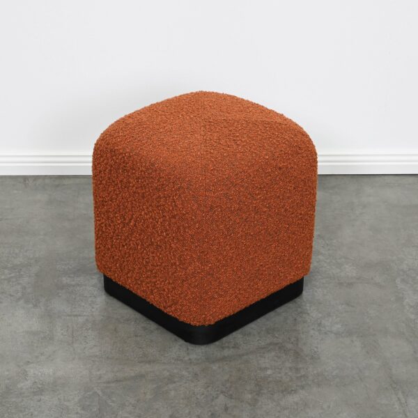 Minimalistic cubic ottoman with soft edges fully upholstered in brick rust boucle on a matte black metal base