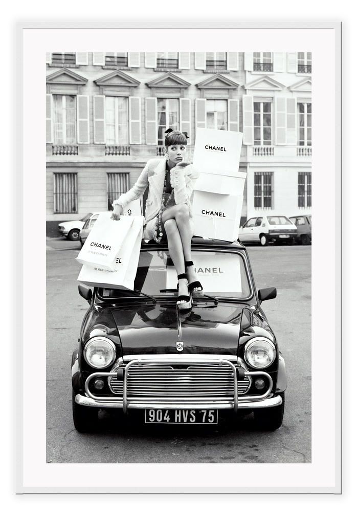 A black and white vintage fashion wall art with a lady holding Chanel shopping bags riding on vintage car Fiat in European city. 