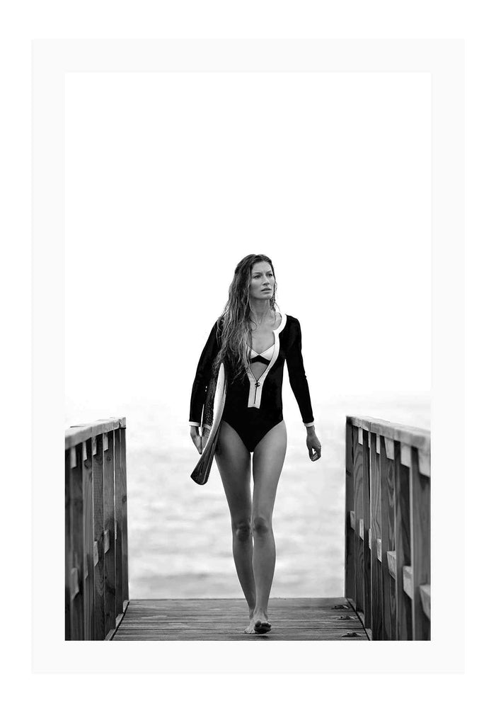 A black and white fashion wall art with Brazilian model Gisele Bundchen holding a surf board walking on timber wharf.
