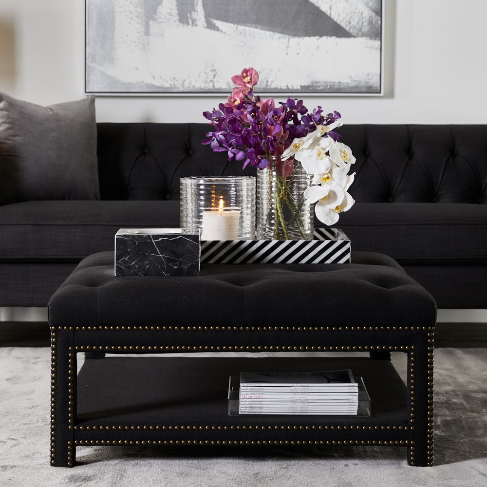 Square shaped ottoman fully upholstered in black linen with tufting detailing on seat cushion and bronze stud detailing on base and legs