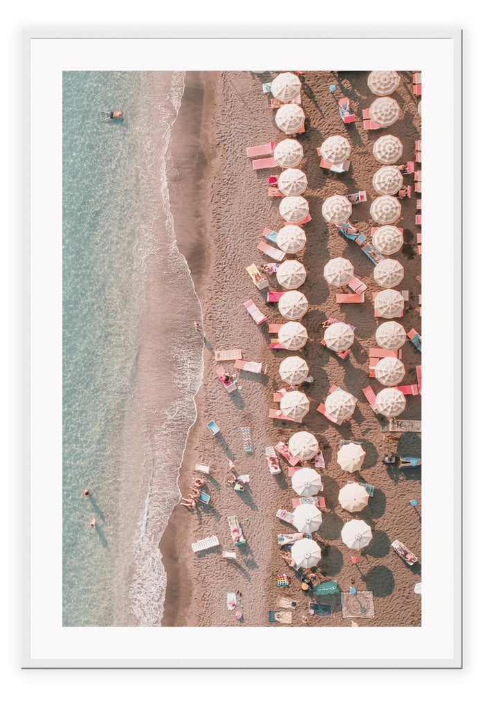 Coastal style photography beach print with pink umbrellas and chairs on the sand in front of the water from a birds eye view.